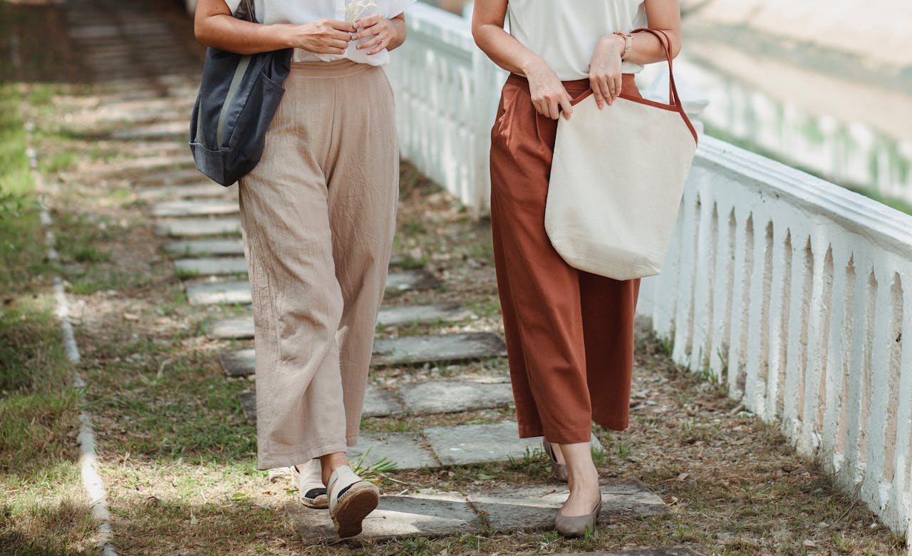 Crop unrecognizable girlfriends in casual wear with bags strolling on fenced walkway in town in summer
