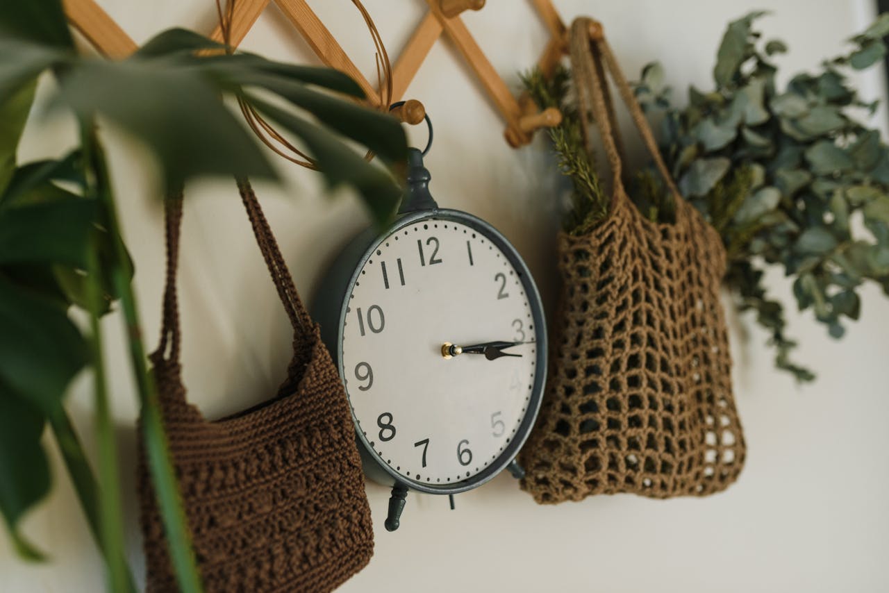 An artistic arrangement featuring a classic clock, crocheted bags, and eucalyptus leaves on a rustic wooden rack.