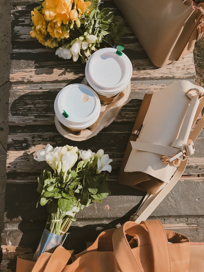 contact-img Flat lay of coffee cups, a bouquet of flowers, and bags on a rustic wooden table under sunlight.
