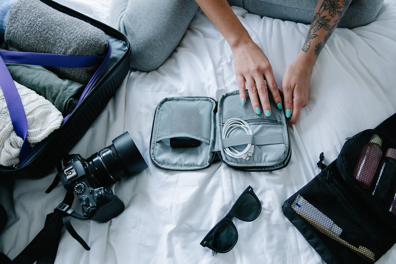 services-04 Woman with tattoos packs electronics, camera, and sunglasses in a suitcase on a bed.